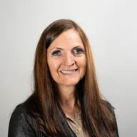 A woman smiling, wearing a dark jacket, in front of a plain light gray background.